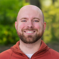 Smiling man, perhaps Blaine Green himself, with a beard and bald head, dons an orange zip-up sweater against a backdrop of blurred greenery.